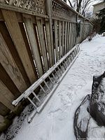 Full length side view of the aluminum extension ladder resting on the snowy ground beside a wooden fence.