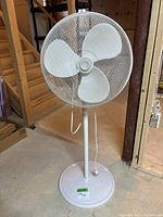 Full view of the white 3 blade oscillating floor fan on a round base in a basement setting.
