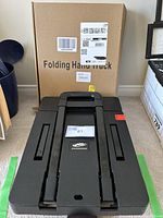 Folded black Spacekeeper folding hand truck with original packaging box behind it, showing compact state and product label.