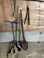 Assorted garden tools leaning against a wall and stacked on the floor. Includes shovel, pruning shears, hoe, cultivator, weed puller and two pairs of small hand shears.