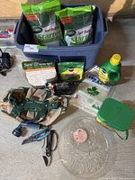 Overview of garden lot with lawn food bags, grass seed bags, garden tote bag, clippers, pruners, twine, hose nozzle, plastic lawn bags, and plant trays in blue storage bin missing lid.
