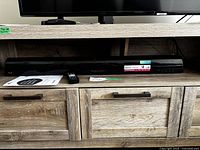 Front view of black LG soundbar placed on wooden shelf, accompanied by remote and user manual