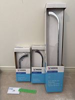 Three boxed Moen Zarina bathroom hardware items including a 24 inch towel bar, mini towel bar, and single post paper holder shown standing against a wall.