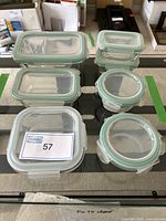 Seven Glasslock glass food storage containers with lids displayed on a black surface. Containers vary in size and shape, both rectangular and round, all with light teal locking lids.