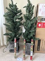 Two green artificial porch trees, pre-lit, approximately 3.5 feet tall, standing in gray wooden-style square bases, two clear tubes of assorted shatter-resistant ornaments placed in front.