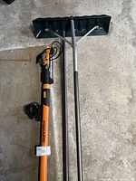 Fiskars brand orange and black electric pruning tool with cord alongside a metal snow shovel with a wide dark green blade, both laid on concrete floor.