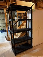 Black Keter stackable plastic shelving unit with five shelves in basement setting next to white cabinet and pumice steps.