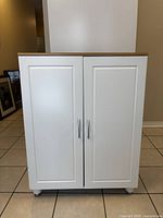 Front view of a white storage cabinet with two doors and silver metal handles, standing on a tiled floor.