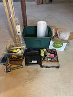 Wide shot showing green plastic bin with roll of bubble wrap, black paint tray, cardboard box with woven protective mats, multiple rolls of tape and tape dispensers, plastic containers with painter supplies and safety eyewear, and a metal caulking gun.