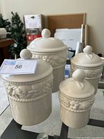 Four cream-colored ceramic canisters with embossed fruit designs and rounded knob lids displayed on glass table.
