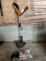 Full side view of the Worx weed trimmer leaning against garage wall, showing handle, shaft, trimming head, charger and plastic bag with parts on floor.