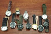 Photo showing 10 assorted pre-owned men's wristwatches laid out on a wooden surface, varying straps and dial colors visible