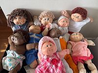 All nine vintage Cabbage Patch Kids dolls arranged on floor showing different skin tones, clothes and hair styles, including one doll with a pink plaid dress and pom-poms.