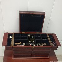 Jewelry box open showing compartments with assorted costume jewelry including bracelets, brooches, earrings, chains, and pins.