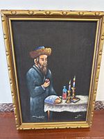 Full view of framed oil painting depicting a Jewish man holding an apple beside a table with ritual Jewish objects including candles, challah bread, kiddush cup, and prayer book on black background.