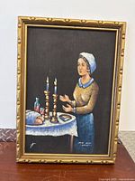Full view of framed oil painting depicting Jewish woman lighting Shabbat candles at a table with ritual objects including challah bread and prayer book, signature visible at bottom.
