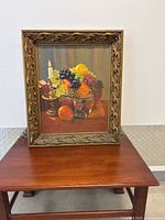 Full frontal image of framed oil painting showing fruit still life on pedestal with candle and books.