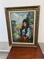 Full view of the framed oil portrait of a woman standing with a natural outdoor background; frame is large with a pattern design.