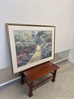 Framed picture leaning against a wall on a small wooden bench, showing full frame and part of the artwork.