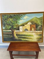 Full view of the framed oil painting showing the landscape scene with a stone building, tree, and mountains.