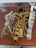 Top-down view of a wooden surface with seven different decorative belts laid out, showing variety in material and style including chains, fabric, rhinestones, and patterned buckles.