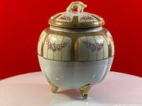 Photo showing entire white porcelain footed biscuit jar decorated with hand painted pink flowers and gold trim, against a red background.
