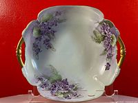 Antique porcelain rectangular tray with hand painted purple flowers and gold-tone bamboo style handles, displayed on clear stand against red background.