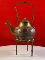 Brass tea pot with floral decorations on a brass stand, shown against red background