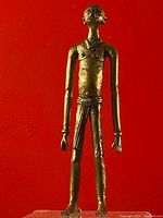 Front view of the brass man figurine with elongated limbs and textured surface against a red background.