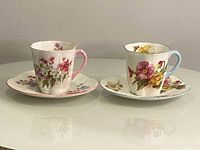 Front view of two floral teacups each on their respective matching saucers showing distinct pink and blue handle designs.