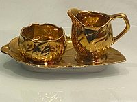 Close-up of the three-piece set including cream pitcher, sugar bowl with lid, atop the matching leaf-shaped dish tray in reflective gold-tone ceramic.