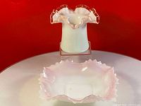 Both the milk glass candy dish and the milk glass hat vase positioned together on a round glass surface, showing white opaque glass with pink ruffled edges against a red background.