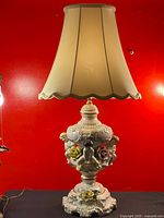 Full view of the vintage Capodimonte porcelain lamp with cream fabric lampshade and ornate base decorated with flowers and cherubs, set against a red background.