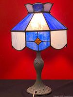 Full view of the vintage table lamp illuminated, showing the stained glass shade in blue and white with amber diamond accents, and ornate metal base.