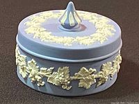 Side view of the round blue Jasperware trinket box with raised white grapevine relief decoration and lid finial.