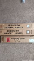 Photo of cardboard boxes labeled Brightshow Laundry Hamper (two visible) and Accordion Wall Mounted Drying Rack, placed on carpet.