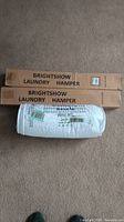 Photo of two boxed Brightshow laundry hampers and a white twin quilt set in original packaging on carpeted floor.