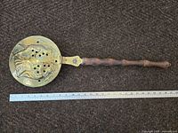 Full image of antique brass bed warmer with wooden handle and metal embossed pan, placed next to metal ruler for size reference