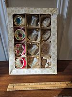 Top view of all ornaments in a divided cardboard box with snowflake design edge and ruler for scale showing 11 ornaments total.