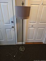 Full view of the antique brass floor lamp with beige fabric drum shade standing on a carpeted floor next to a white door. Power cord coiled at base.