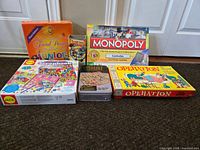 Photo showing the lot of 6 board and activity games arranged on the floor in front of a white door, items include Monopoly Canada, Operation, Junior Trivial Pursuit, Alex Scrapbook Kit, Chutes and Ladders, and a puzzle