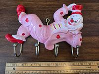 Pink chalkware clown coat rack with red accents and four metal hooks shown on a wooden surface with ruler for scale.
