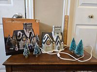 Ceramic Christmas village church with light cord and four small artificial snowy pine trees displayed on a wooden surface alongside original box.