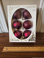 Six cranberry colored round glass ornaments in original white packaging with gold accents from The Victoria Collection, front view.