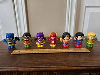 Seven Fisher Price Little People DC Comics superhero figures lined up on a wooden surface with a ruler for scale. Characters included are Batman, Robin, Batgirl, Flash, Wonder Woman, Superman, and Green Lantern.