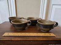 Four brown and beige speckled pottery mugs with dark horizontal banding around middle, shown with ruler for scale