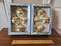 Two open blue boxes with four pearl white glass ornaments each, decorated with gold glitter trim and leaf motifs, arranged on a surface with a ruler for scale.