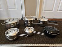 Set of stainless steel pots, saucepans, frying pan with lids shown from front with ruler for scale