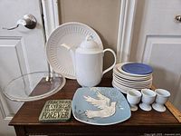 Wide shot of dishware lot including coffee pot, large plate, blue rimmed plates, bowls, egg cups, glass pie dish, and decorative plaques.