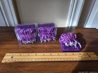 Two sets of purple string lights coiled inside clear plastic boxes, resting on a wooden surface next to a ruler for size reference.